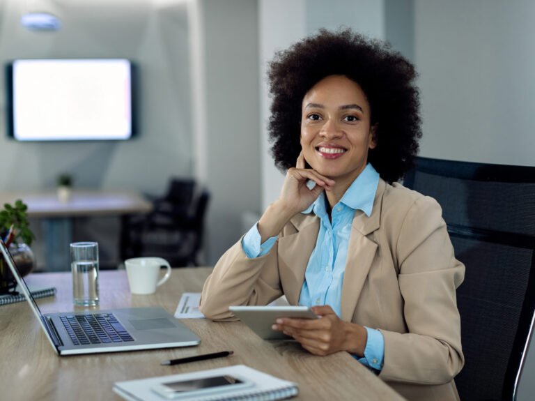 Happy black entrepreneur working on wireless technology at her office desk and looking at camera.